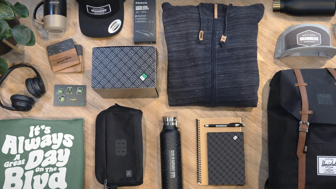 Diverse collection of branded swag. Trendy high-quality products, including Skullcandy wireless earbuds, Tentree hoodie, Herschel backpack, water bottles, notebooks, hats, and packaging for effective brand recognition and loyalty.
