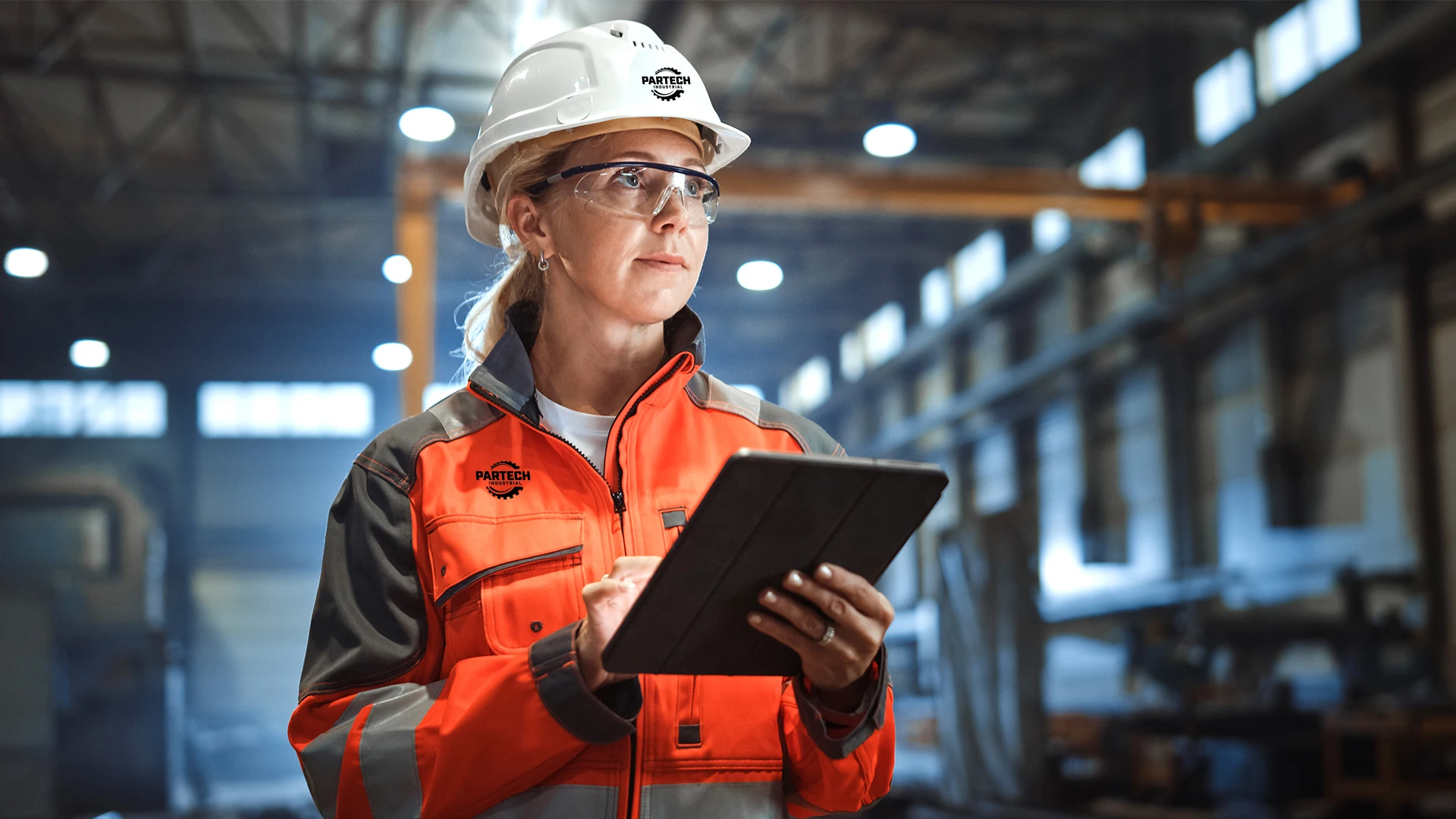 Empowered female manufacturing worker in branded swag holding an iPad, wearing a hard hat, safety glasses, and orange safety jacket in an industrial warehouse. High-quality safety gear showcased in a modern manufacturing setting.