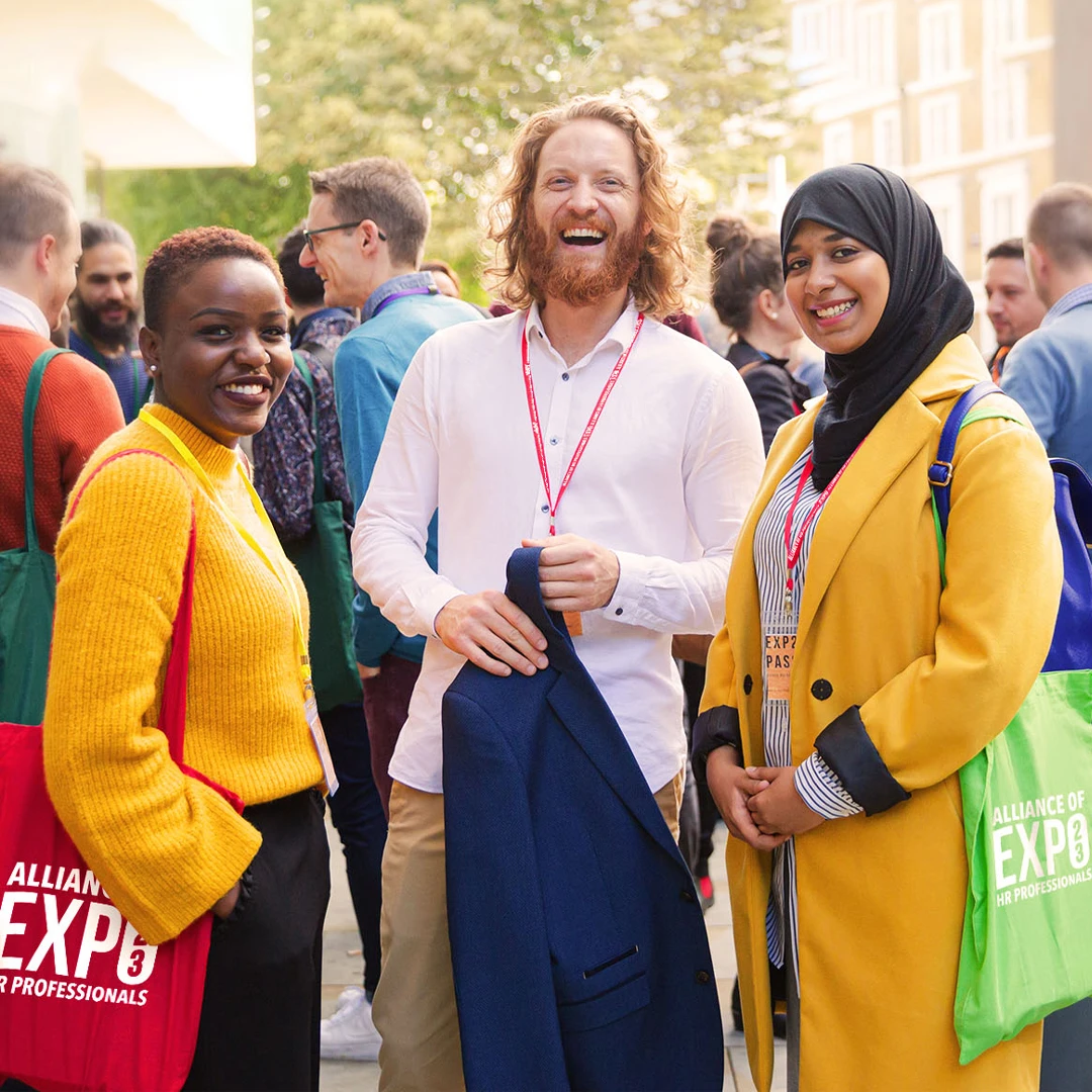 Three engaged member conference attendees wearing event swag, including branded tote bags and lanyards from their membership organization. Great examples of custom branded merch and promotional products that foster a strong sense of community.