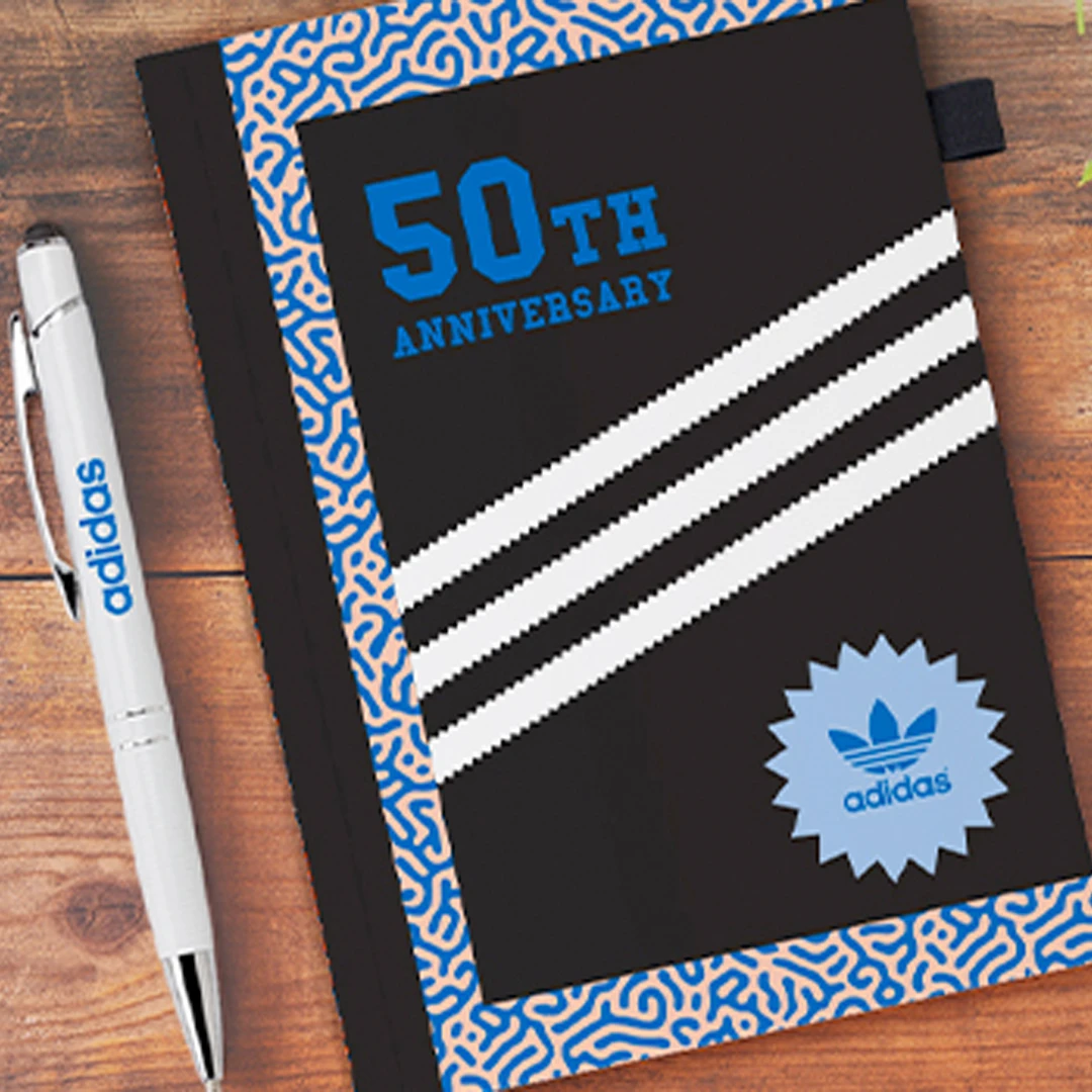 A photo of a small notebook and pen branded with the Adidas logo and 50th anniversary logo on a wood table.