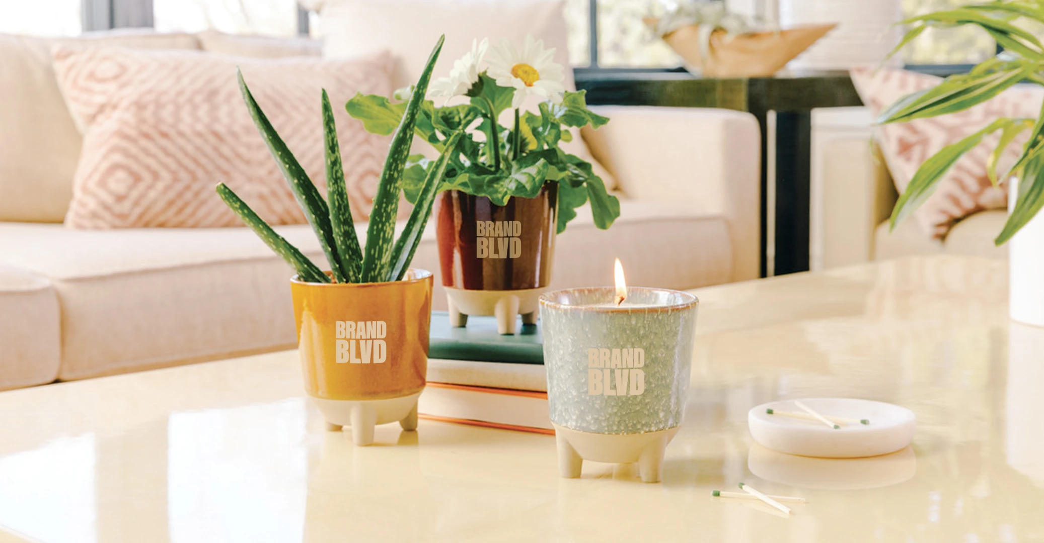 Two potted plants and a candle branded with the Brand Blvd logo, sitting on a coffee table in a living area in daylight.