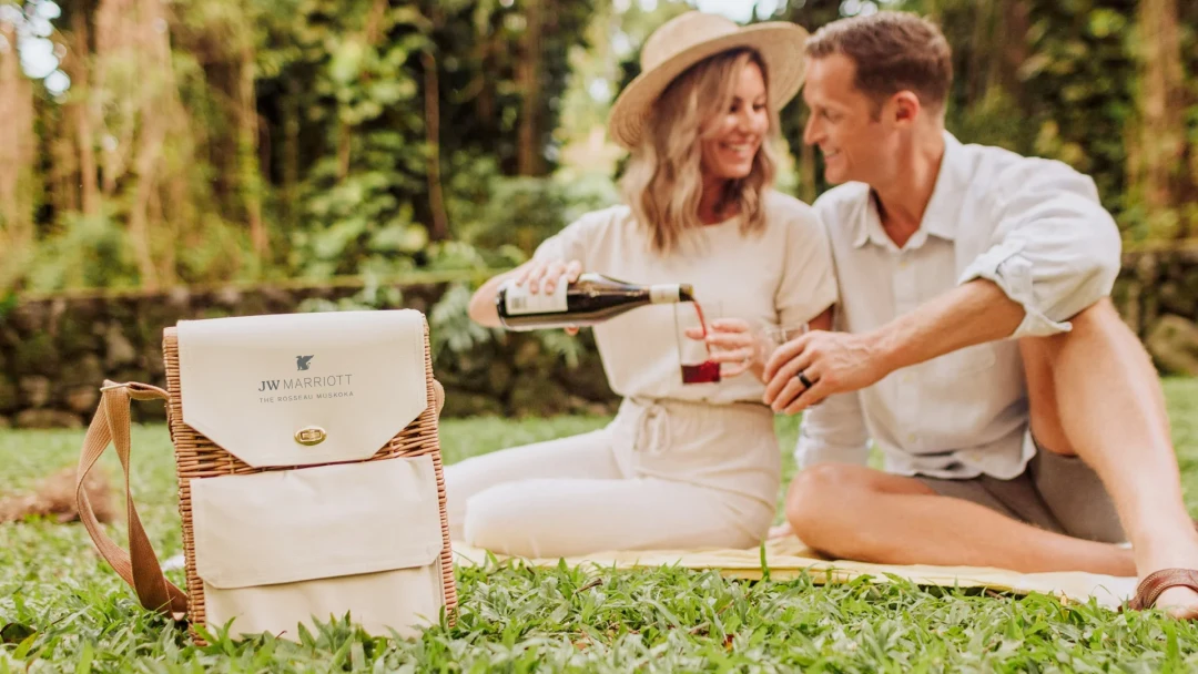 A couple enjoying a high-quality welcome gift wicker picnic basket, branded with the JW Marriott - The Rosseau Muskoka logo. An example of quality branded merch for hospitality guest engagement from Brand Blvd.