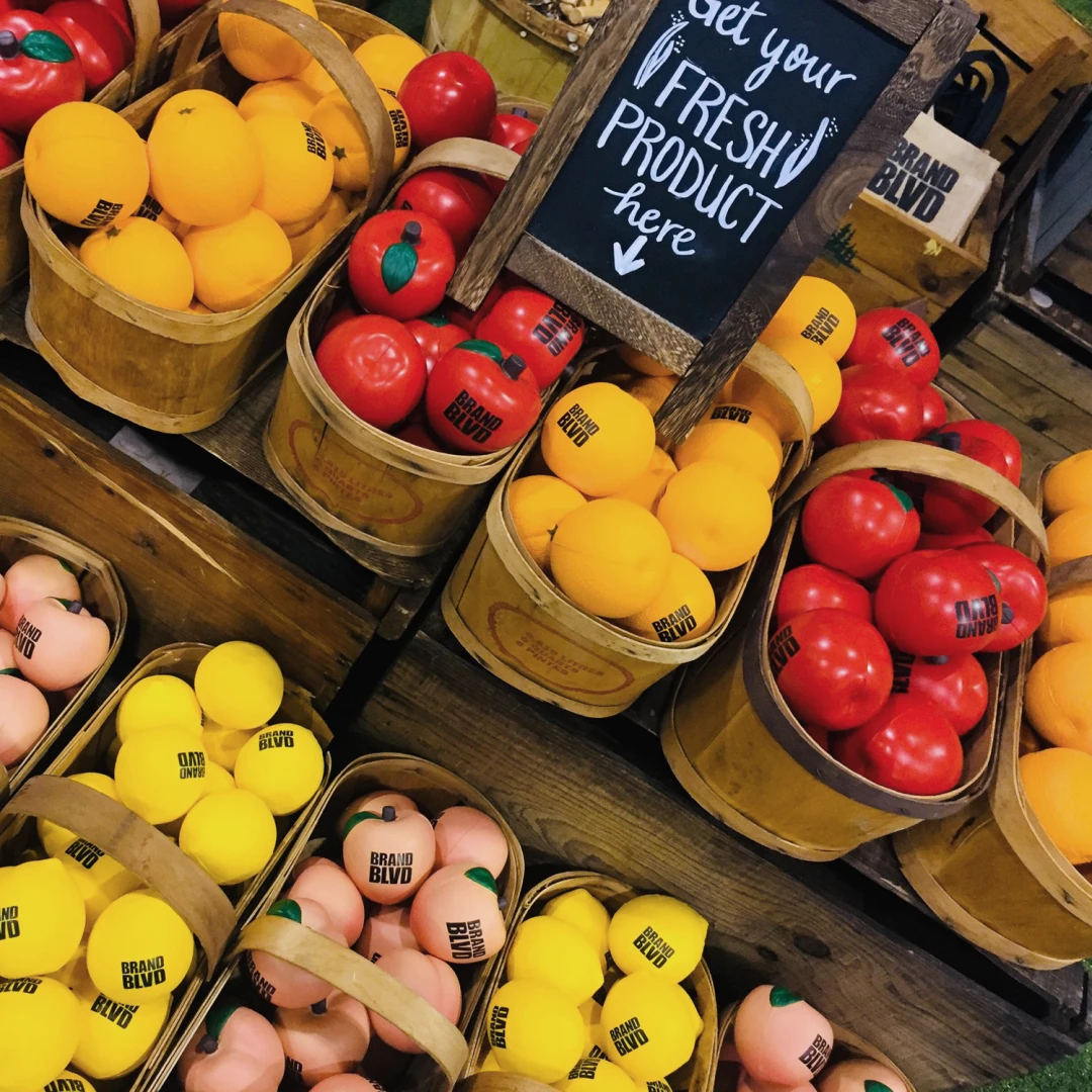 Strategically design your booth and give away free branded promotional items at your trade show booth as part of your event marketing strategy to attract people and create a buzz, such as these branded stress balls that look like fruit in a market stand. 