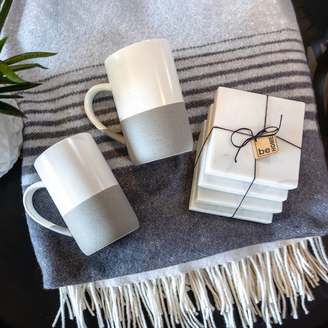 A photo of the Home Luxe curated swag kit which includes a fringed grey striped blanket, natural toned ceramic speckled mug and a set of 4 white marble coasters.