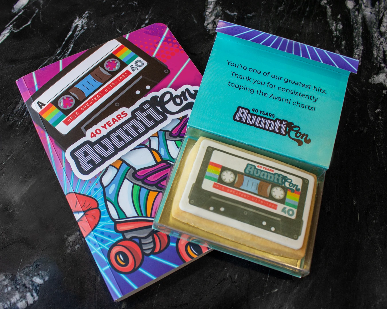 A photo of a notebook with colourful 80s design of rollerskates and a cassette and the Avanati Con 40 years logo. And a sugar cookie postcard with a cassette printed on the icing with a thank you message on a black marble table.