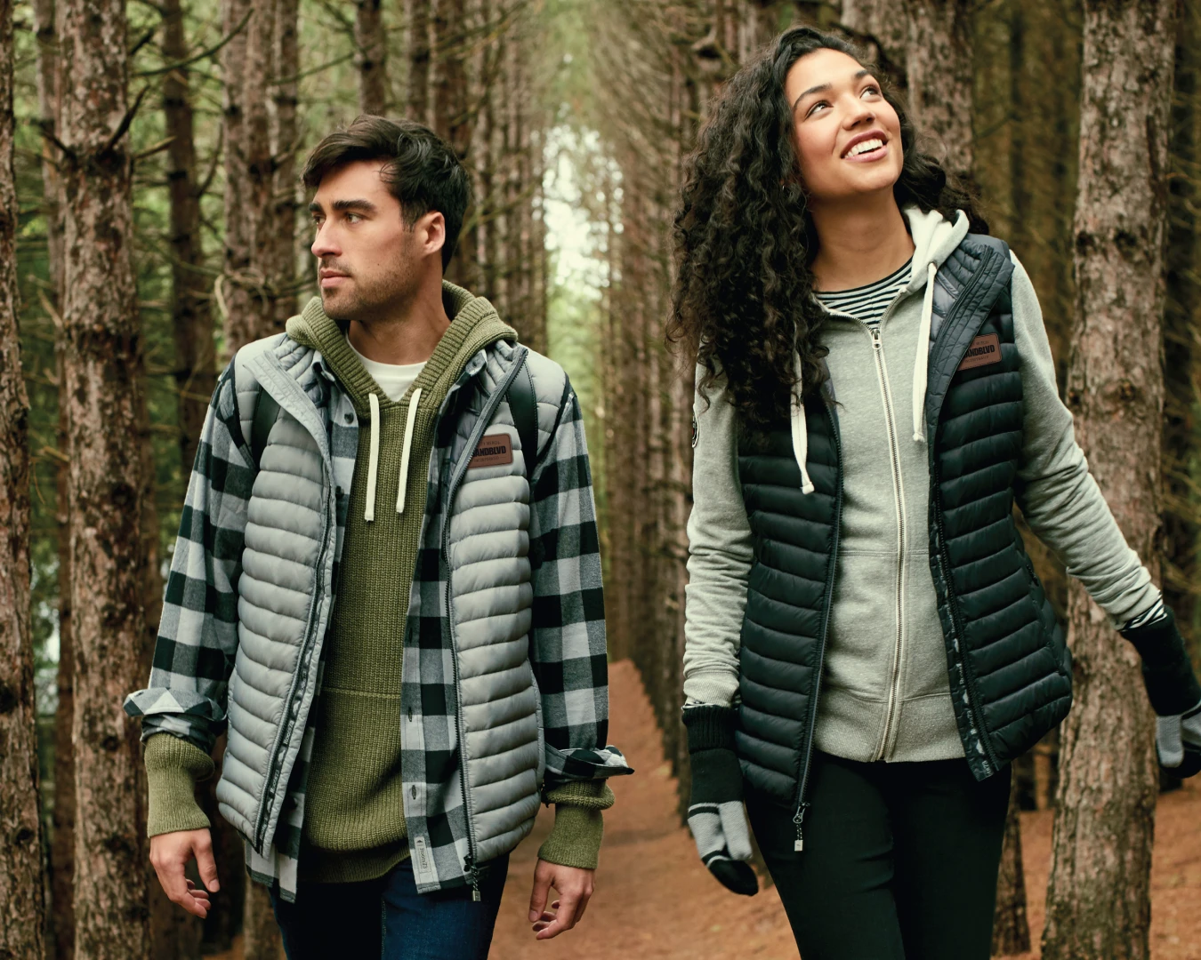 A photo of a man and woman in branded Roots apparel, walking through the woods smiling.