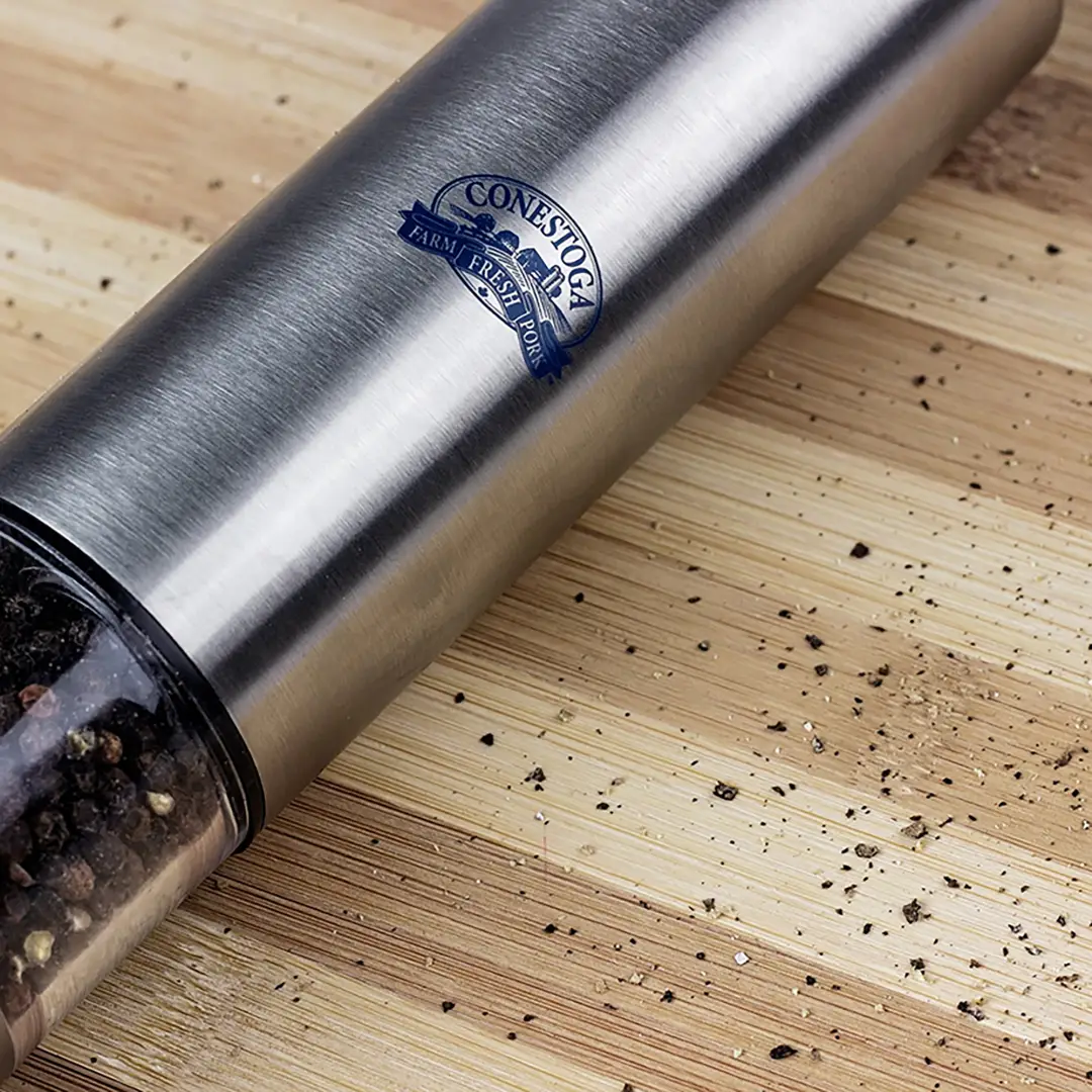 A photo of a chrome and glass pepper mill with the Conestoga Meats logo imprinted in navy blue, on top of a wood cutting board with ground pepper flakes spread around the mill. 