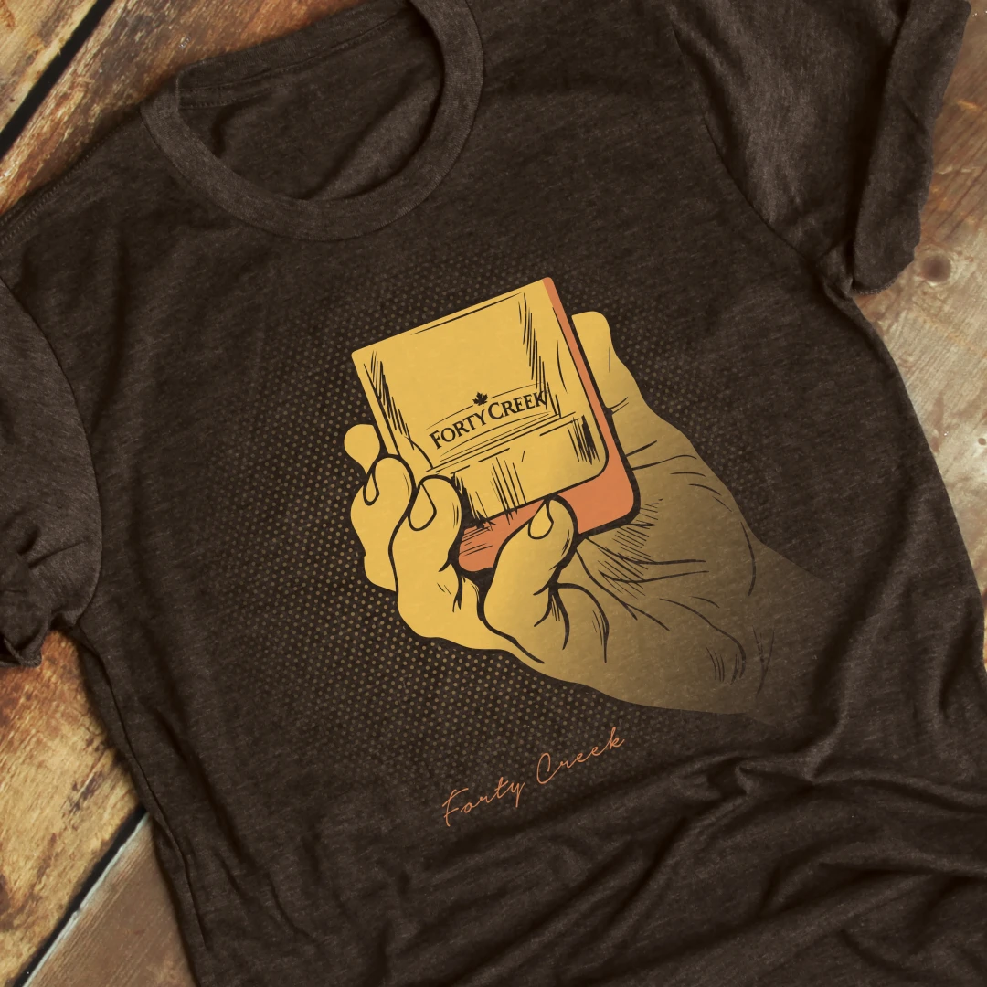 A photo of a brown t-shirt with a Forty Creek Whisky printed design of a yellow and orange hand holding a whisky glass, flat layed on a wood background.
