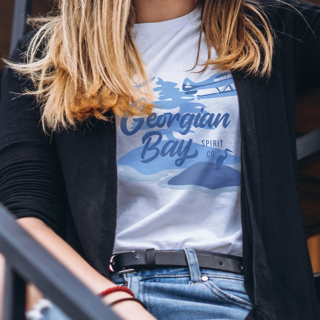 A close up photo of a woman with blonde hair wearing a white t-shirt with a Georgian Bay Spirit Company graphic design printed on it, light blue jeans and a black cardigan sweater.