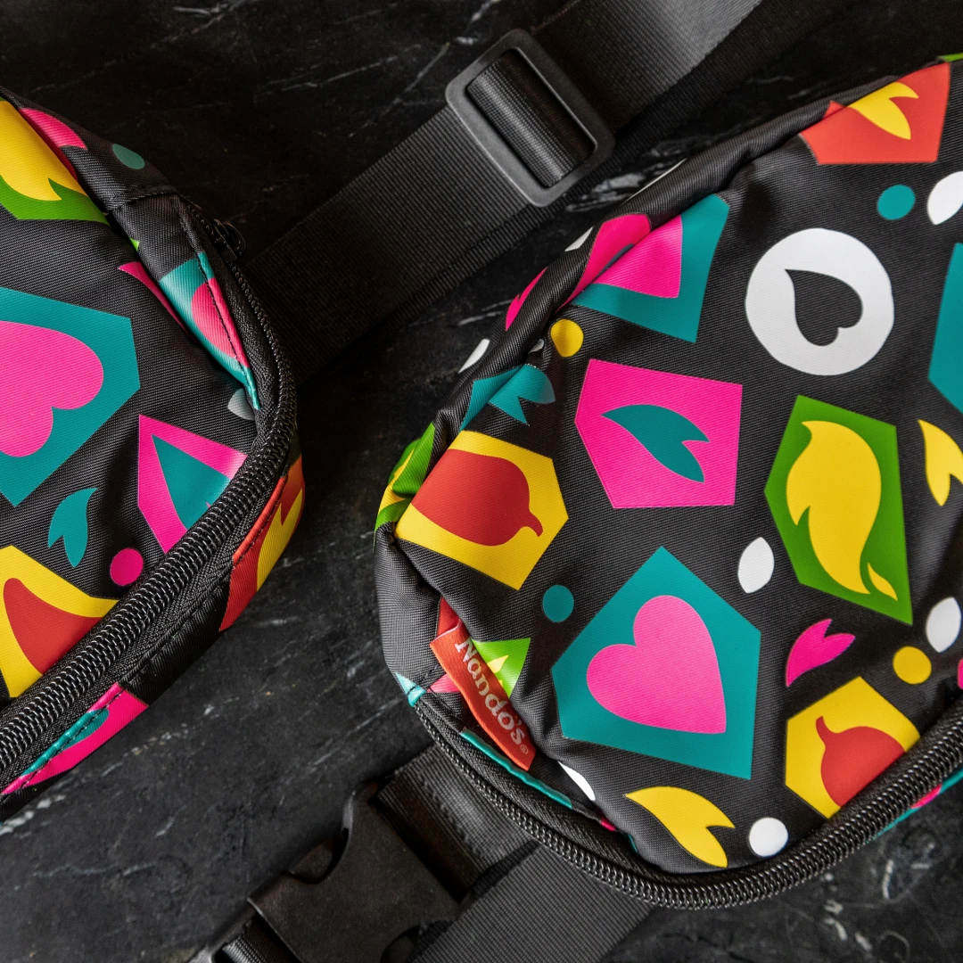 A photo of two black fanny packs with colourful shapes printed and a red woven label with the Nando's logo, on a black marble background.