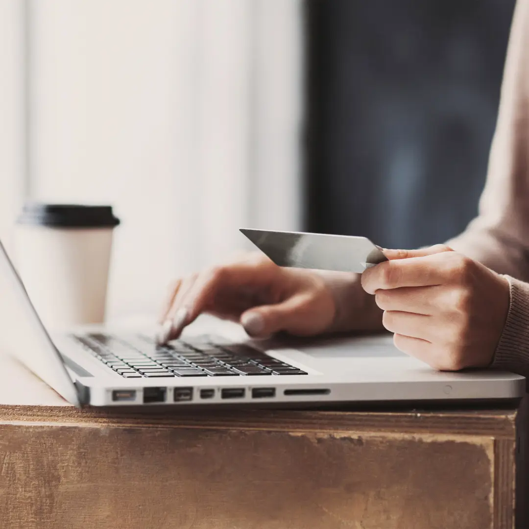 A photo of a person on laptop, holding their credit card making a payment online with a coffee cup on the desk.